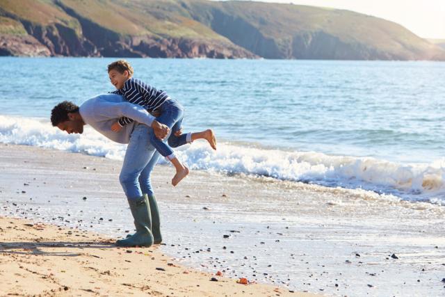 father giving his son a piggyback as they walk along a beach together. UK