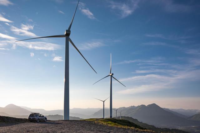 Five wind turbines on a mountain range with a silver car parked next to one