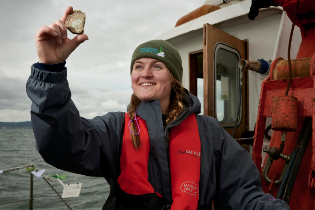 Emmy smiling and holding up a native oyster into the air.