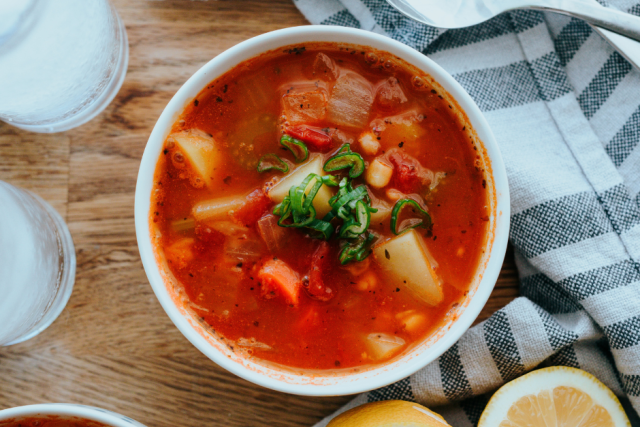 Two white ceramic bowls of minestrone soup, placed on top of a tea towel. Beside the soup is two spoons, glasses of water and slices lemon.