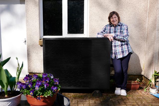 Lesley stood proudly with her heat pump. The heat pump is beside the outside of her home and surrounded by plant pots with bright purple flowers.