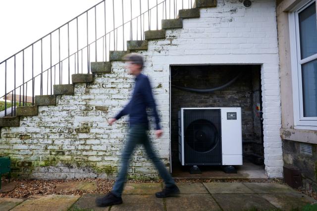 Huw walking past his heat pump outside their Edinburgh home. They're both smiling at the camera.