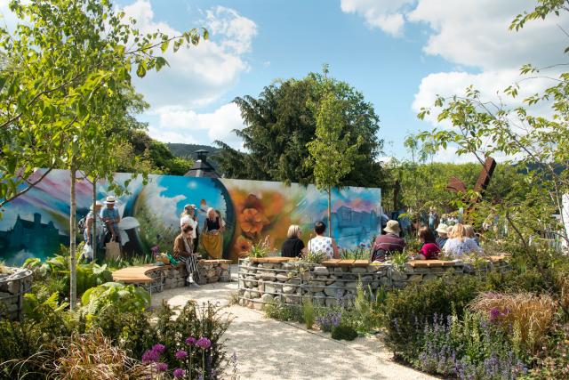 Greening Maindee garden in Malvern Show. Stone walls encompass a weaving gravel path, with young trees and a variety of shrubs and plants. People can be seen sitting on parts of the wall that are designed as benches and a small crowd is standing looking at a large colourful mural at the back, which features vibrant flowers, buildings and bright blue skies.