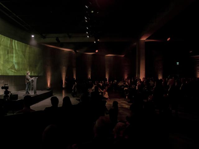 A large, dark room with a speaker in front of a green background addressing a large, seated audience