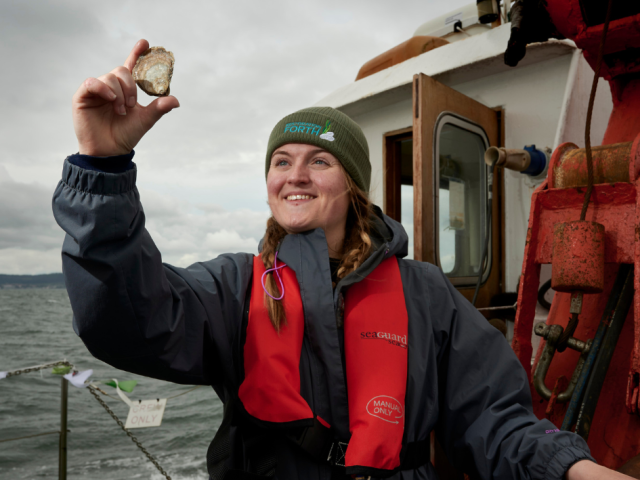 Emmy smiling and holding up a native oyster into the air.