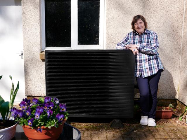 Lesley stood proudly with her heat pump. The heat pump is beside the outside of her home and surrounded by plant pots with bright purple flowers.