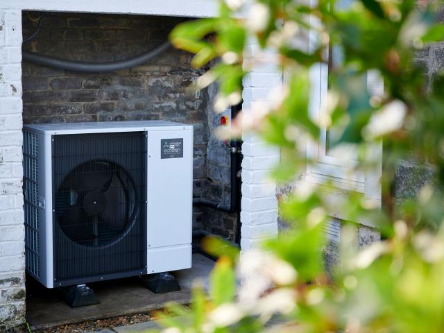 A heat pump on the side of an Edinburgh home. Green foliage in the forefront.