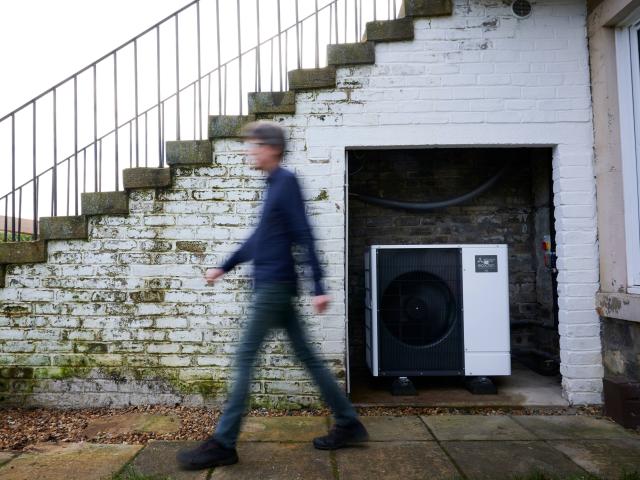 Huw walking past his heat pump outside their Edinburgh home. They're both smiling at the camera.