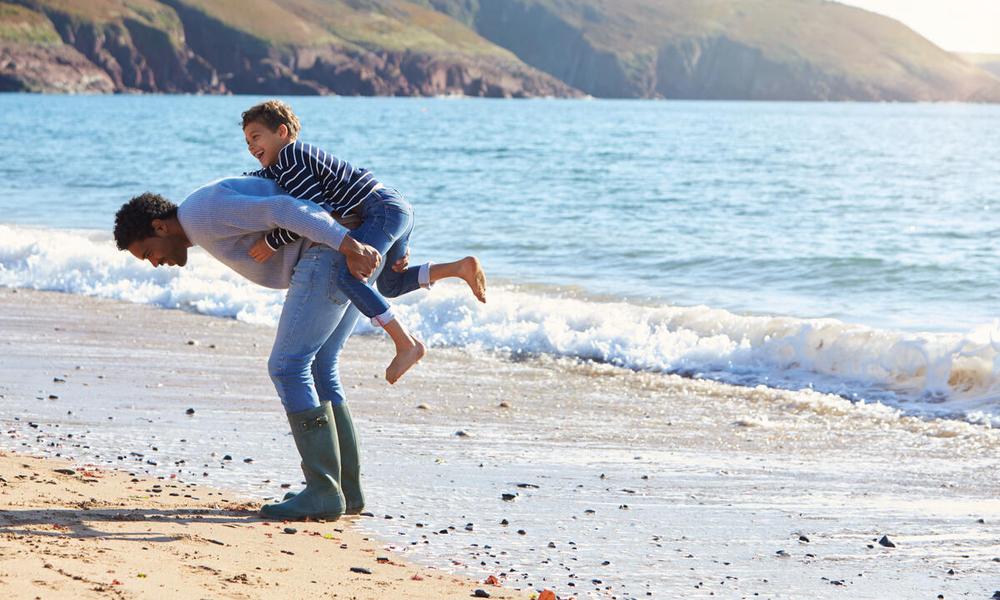 father giving his son a piggyback as they walk along a beach together. UK
