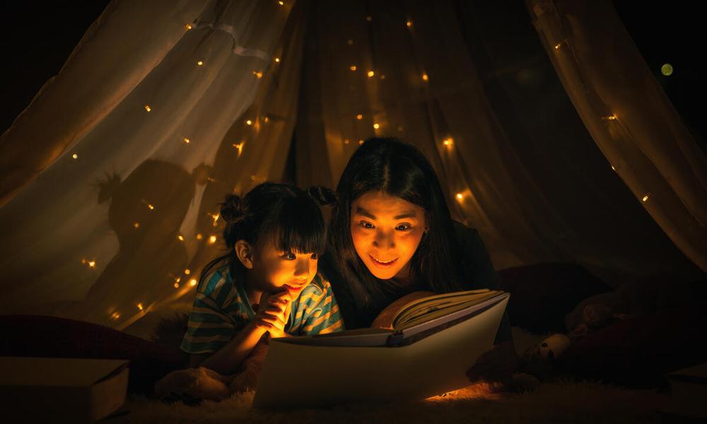 Mother and daughter read a bedtime story under a made up tent in their bedroom. 