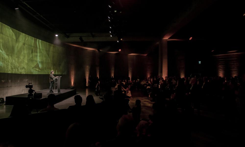 A large, dark room with a speaker in front of a green background addressing a large, seated audience