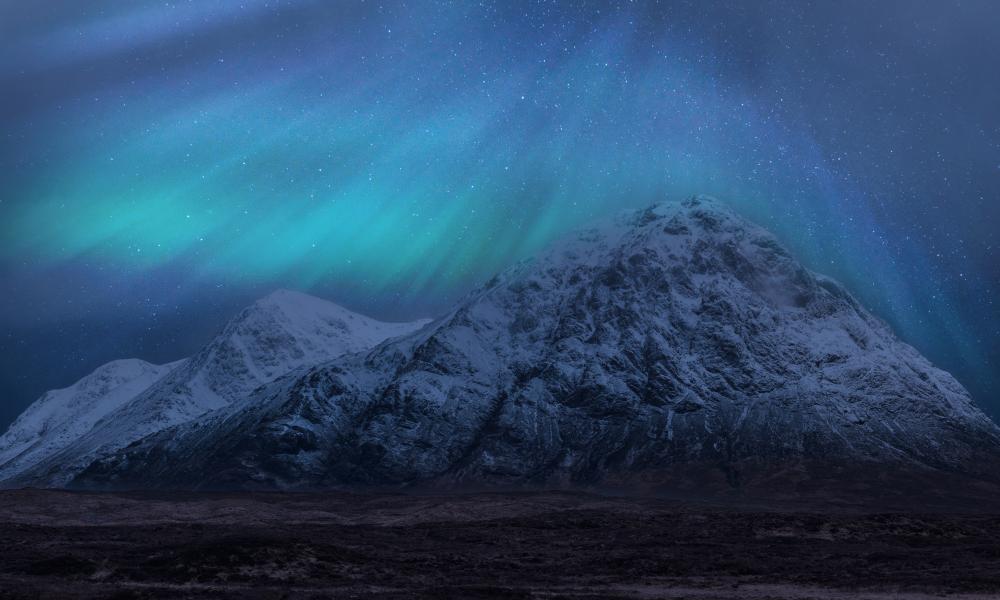 Bright blue and green northern lights dance across the sky above snow capped mountains in the Scottish Highlands.