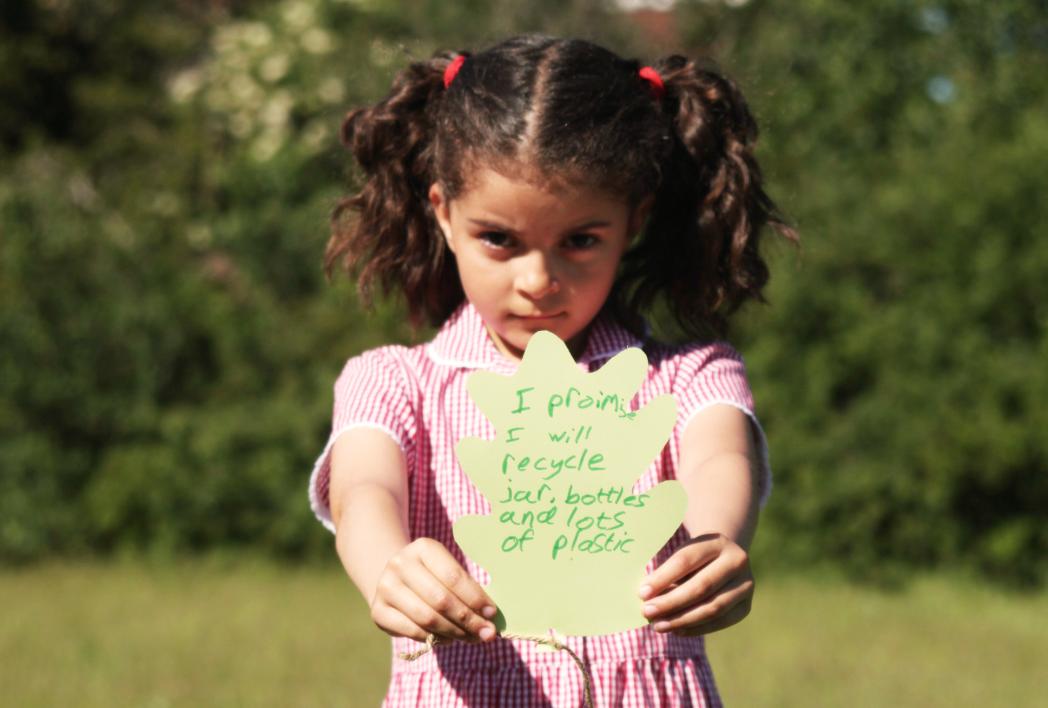 A girl in school uniform holds a card leaf towards the camera. On it is written "I promise to recycle jars, bottles and lots of plastic."
