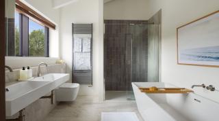 Omaze Cornwall house bathroom, with a white modern bath, two with sinks with metal taps, toilet, heated towel rail and grey tiled shower with glass doors. A picture of the ocean on one wall and a window on the exterior wall.