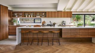 Modern kitchen area with wooden floors and cupboards with four stools lined up along a bar and a large window looking out onto a green garden area.