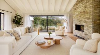 Omaze Cornwall house lounge, cream coloured long sofa and two chairs, with three wooden coffee tables and a built in fire place with stone exterior. Looking though on to two large windows to the outside.