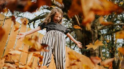 Autumn leaves scatter, falling towards the camera, surrounding a girl as she laughs after throwing a large handful into the air. Depicts a carefree childhood, and autumn time. 