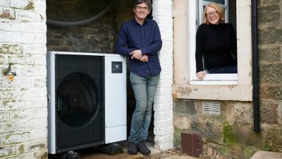 Huw and Kristen stood beside their heat pump outside their Edinburgh home. They're both smiling at the camera.