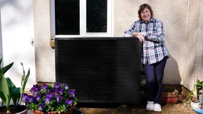 Lesley stood beside her heat pump outside her Grandtully home. She is smiling at the camera.