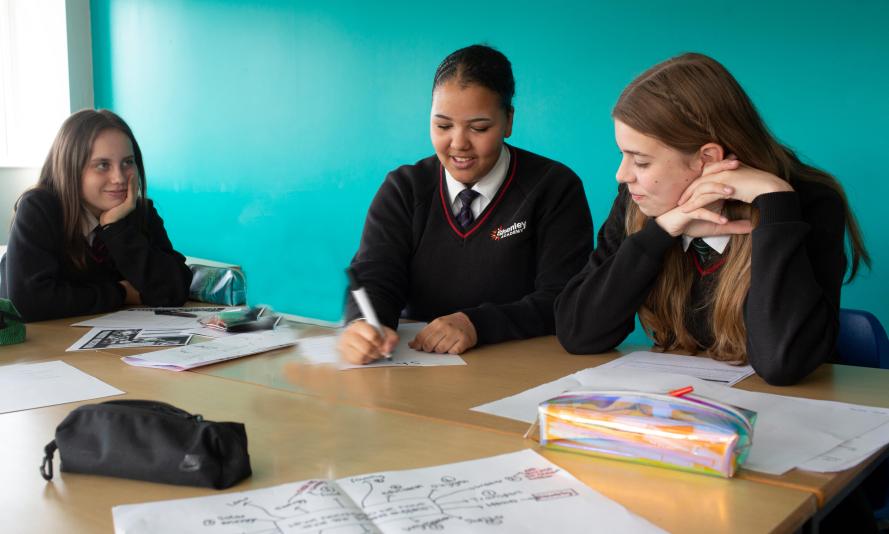Three school pupils sat at around a table writing on paper Three school pupils sat at around a table writing on paper