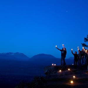 A group of men and women gathering on a clifftop on Vancouver Island against silhouetted trees and a twilight sky looking toward the sky and forested mountain scenery