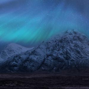 Bright blue and green northern lights dance across the sky above snow capped mountains in the Scottish Highlands.